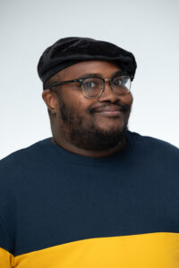 Photo of a Black man wearing a black velvet beret and a colour-block long-sleeved shirt.